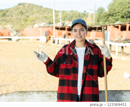 Smiling skilled young woman in plaid shirt smiling at camera while holding working tool on horse ranch in spring Smiling skilled young woman in plaid shirt smiling at camera while holding working tool on horse ranch in spring 123369451