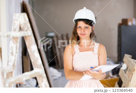 Girl in beige dress and protective helmet notes in document the completed stages of repair work in the cottage 123369540