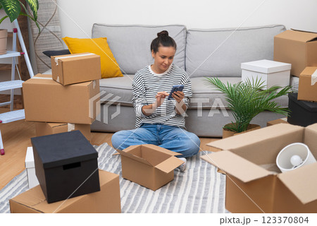 Young woman unpacks on moving day, stays connected with her phone in her new home, marking a new chapter Young woman unpacks on moving day, stays connected with her phone in her new home, marking a new chapter 123370804