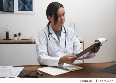Female doctor diagnoses patient studying documents on clipboard medical specialist reads test results and makes notes sitting at table in light office 123370827