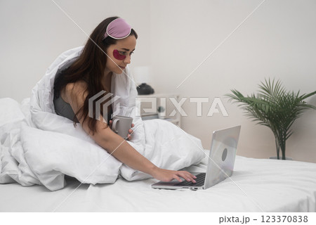 Bed becomes her workspace as she wears eye patches, sips coffee, and tackles online work and remote studies on her laptop from home.  123370838