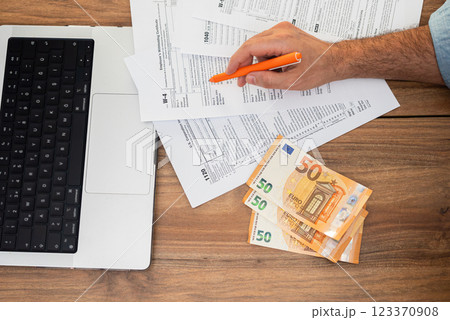 Male entrepreneur points with pen at tax forms carefully examining filling requirements sitting at wooden workplace with euro banknotes and corporate laptop Male entrepreneur points with pen at tax forms carefully examining filling requirements sitting at wooden workplace with euro banknotes and corporate laptop 123370908