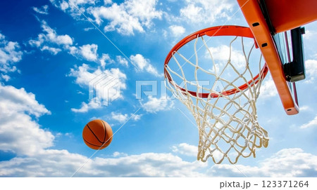 Basketball ball flying through the air towards the hoop against a backdrop of bright blue sky and white clouds, capturing the essence of outdoor sports and athletic competition 123371264
