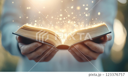 Two hands holding open book glowing softly in sunlight, radiating magic 123376757