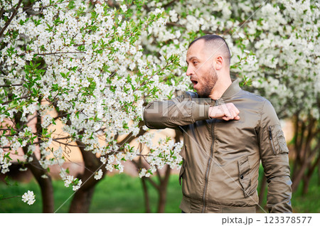Man allergic suffering from seasonal allergy at spring in blossoming garden at springtime. Bearded young man sneezing and having runny nose in front of blooming tree. Spring allergy concept. Man allergic suffering from seasonal allergy at spring in blossoming garden at springtime. Bearded young man sneezing and having runny nose in front of blooming tree. Spring allergy concept. 123378577