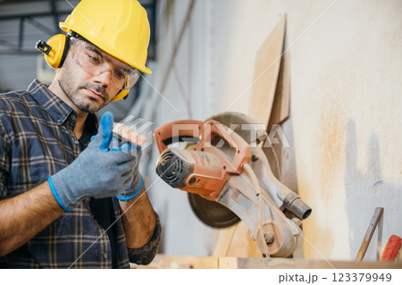 Carpenters man in a yellow hardhat, safety glasses measuring wood for cutting, with a circular saw in the background. Ideal for woodworking and carpentry workshop settings, National Carpenters Day 123379949