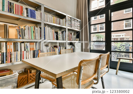 A stylish modern interior featuring an organized bookshelf filled with books, a spacious table, and large windows allowing natural light to brighten the space. 123380002