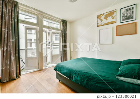 A serene and minimalistic bedroom featuring a green bedspread, large windows, and wooden flooring, creating a tranquil space with ample natural light. 123380021