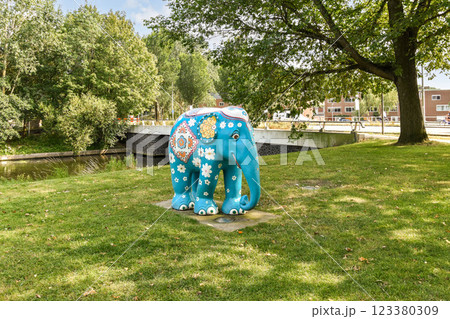 A vibrant blue elephant sculpture adorned with intricate designs stands in a lush green park by a serene waterway, surrounded by trees and buildings. A vibrant blue elephant sculpture adorned with intricate designs stands in a lush green park by a serene waterway, surrounded by trees and buildings. 123380309