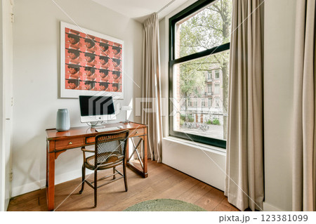 A stylish home office featuring a wooden desk, computer, and large window with curtains that lets in natural light, creating a serene workspace atmosphere. 123381099