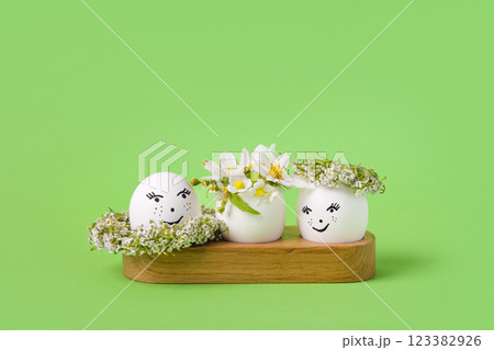 Three white eggs with a cheerful face and spring flowers of pears, strawberries and a white wreath of flowers on a green background. Happy Easter greeting card with a place for text. 123382926