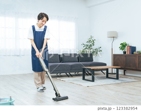 部屋の床に掃除機をかけるエプロンをつけたミドル女性 123382954