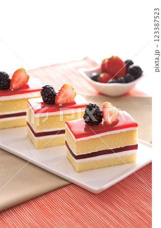Sweet and colorful layered cakes with a glossy red topping, fresh strawberries, and blackberries. The soft sponge, creamy layers, and fruity flavors make it a tasty dessert. 123387523