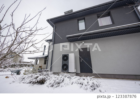 Outdoor heat pump unit mounted on the wall of a modern gray house surrounded by snow-covered bushes and a winter garden setting. Outdoor heat pump unit mounted on the wall of a modern gray house surrounded by snow-covered bushes and a winter garden setting. 123387876