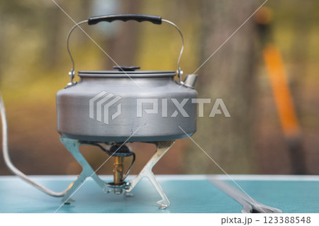 A stainless steel kettle sits on a camping stove, ready for use in a peaceful outdoor environment filled with trees. A stainless steel kettle sits on a camping stove, ready for use in a peaceful outdoor environment filled with trees. 123388548