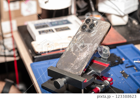 A damaged smartphone with broken glass is placed in a repair stand at a phone service shop, surrounded by tools and equipment. A damaged smartphone with broken glass is placed in a repair stand at a phone service shop, surrounded by tools and equipment. 123388623
