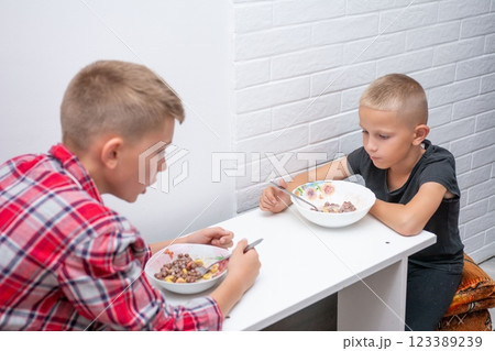 Two boys sit at a white table, enjoying a morning meal of cereal. They are focused on eating. Casual dining scene with white brick wall background. 123389239