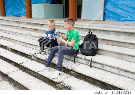 Two young boys sit on outdoor concrete steps, one opening a water bottle, with a backpack beside them, enjoying a break outside. 123389252