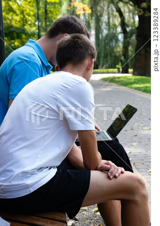 Two students collaborate on a laptop while sitting on a park bench, focusing on studies in an outdoor learning environment. Two students collaborate on a laptop while sitting on a park bench, focusing on studies in an outdoor learning environment. 123389284