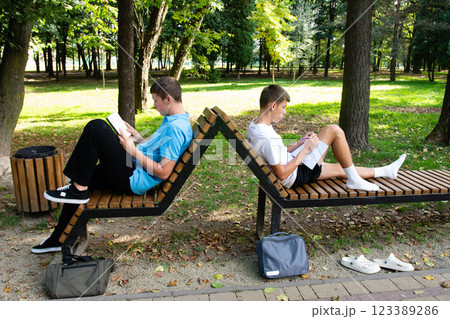 Two young people reading and writing while relaxing on a unique wooden bench in a park surrounded by lush greenery. Two young people reading and writing while relaxing on a unique wooden bench in a park surrounded by lush greenery. 123389286