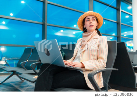 Mid shot of Caucasian elegant young woman is sitting on a chair in the waiting room of airport and working on a laptop. Bottom view. Concept of remote work and travel Mid shot of Caucasian elegant young woman is sitting on a chair in the waiting room of airport and working on a laptop. Bottom view. Concept of remote work and travel 123390830