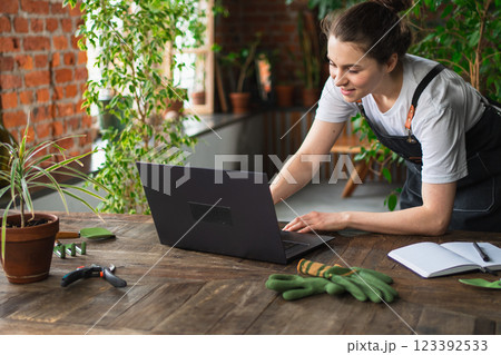 Female florist gardener working in botanical store greenhouse using laptop order plants communicating on Internet with customer. Small business owner working at flower shop. Running of own business Female florist gardener working in botanical store greenhouse using laptop order plants communicating on Internet with customer. Small business owner working at flower shop. Running of own business 123392533