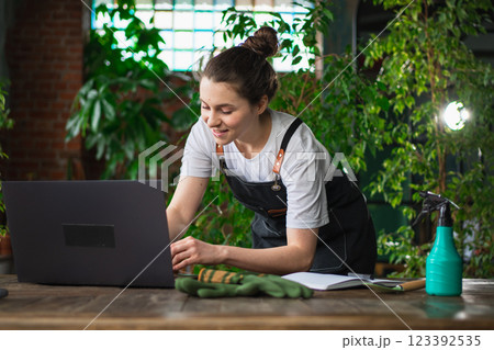 Female florist gardener working in botanical store greenhouse using laptop order plants communicating on Internet with customer. Small business owner working at flower shop. Running of own business Female florist gardener working in botanical store greenhouse using laptop order plants communicating on Internet with customer. Small business owner working at flower shop. Running of own business 123392535