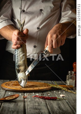 A dedicated chef expertly holds a whole fish while preparing it on a wooden cutting board. The kitchen exudes warmth, with spices and herbs adding to the atmosphere of culinary craftsmanship A dedicated chef expertly holds a whole fish while preparing it on a wooden cutting board. The kitchen exudes warmth, with spices and herbs adding to the atmosphere of culinary craftsmanship 123393711