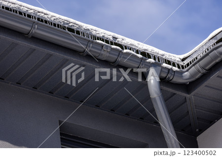 Modern metal gutter system with icicles and snow-covered eaves, highlighting winter conditions and roof drainage against a clear blue sky. Modern metal gutter system with icicles and snow-covered eaves, highlighting winter conditions and roof drainage against a clear blue sky. 123400502