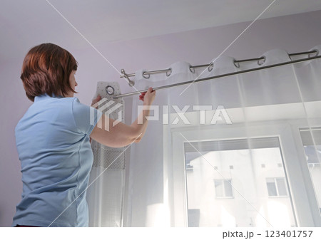 Woman hangs white curtains with metal rings made of natural material with embroidered hearts in kitchen of house. Cleaning and cleanliness. Interior and textiles. Woman hangs white curtains with metal rings made of natural material with embroidered hearts in kitchen of house. Cleaning and cleanliness. Interior and textiles. 123401757