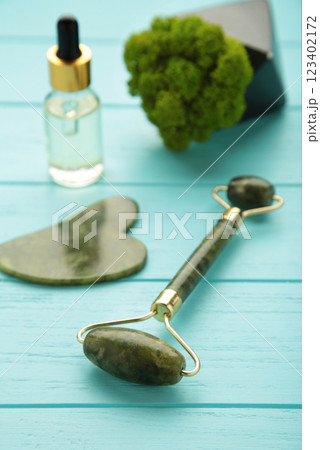 Green jade roller and gua sha stone for facial massage and on blue background. Home beauty and selfcare accessories. Face roller for anti age wrinkle treatment. Top view, flat lay. 123402172