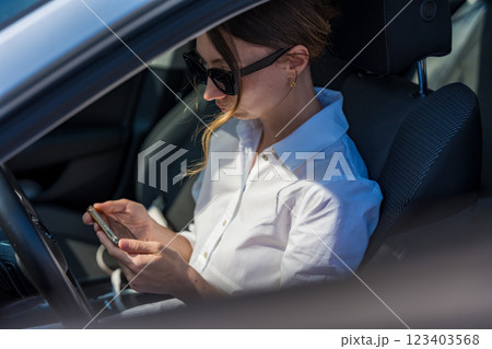 A woman in sunglasses and a white shirt sits in a car, holding a smartphone tilted horizontally.  123403568