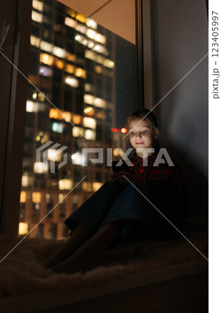 Vertical portrait of pretty little girl browsing smartphone, illuminated in digital light, sitting on windowsill in dark room with soft bokeh light on blurred background creating nighttime ambiance. Vertical portrait of pretty little girl browsing smartphone, illuminated in digital light, sitting on windowsill in dark room with soft bokeh light on blurred background creating nighttime ambiance. 123405997