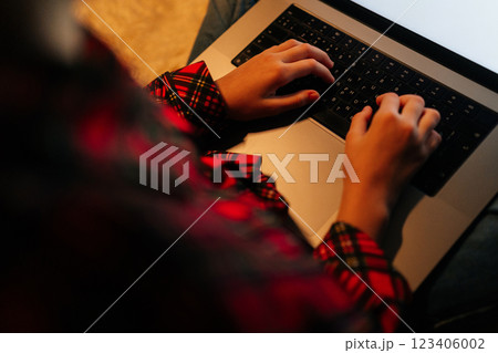 View from shoulder of unrecognizable child girl wearing red and black checkered shirt typing on backlit laptop keyboard sitting on bed with crossed legs in dark room, creating cozy atmosphere. 123406002