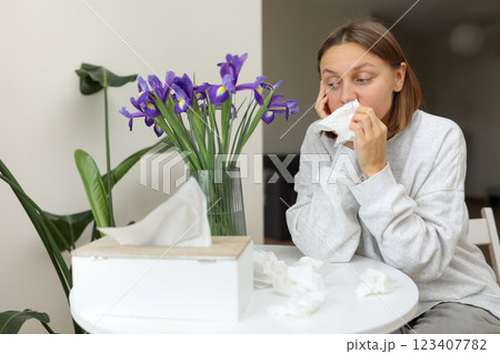 Sick unhealthy allergic young woman covering nose with paper tissue has runny stuffy nose, sneezes from iris flowers pollen at kitchen home. Girl with flu, itching or cough, seasonal allergy,rhinitis. 123407782