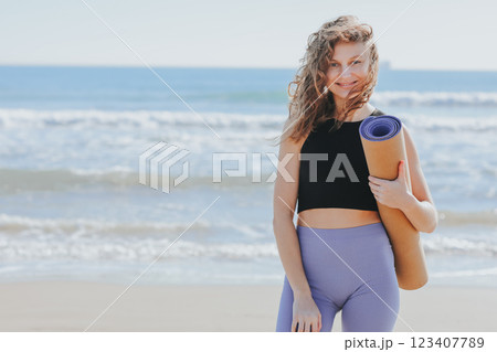 Young woman holds yoga mat, looks at camera and smiles. Lady prepares to workout outdoors. Sport and fitness on seashore. Healthy lifestyle, morning exercises, female wellness and power, healthcare. 123407789