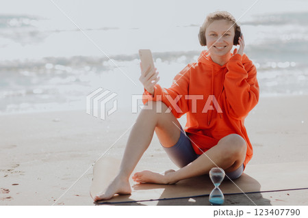 Workout playlist. Happy young caucasian woman with earphones sits on yoga mat outdoors on sea beach, listens to music or browsing internet with smartphone while resting after fitness exercising. 123407790