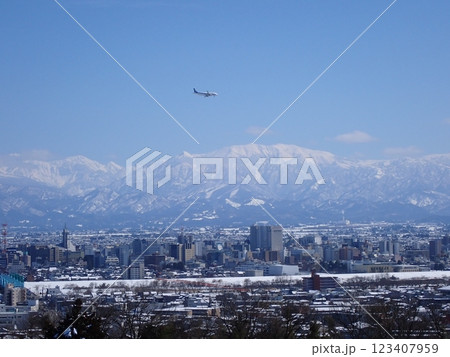 冠雪の北アルプス薬師岳を背景に雪景色の富山市街地と着陸態勢の旅客機 123407959