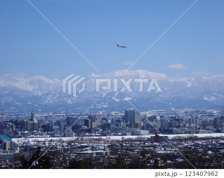 冠雪の北アルプス薬師岳を背景に雪景色の富山市街地と着陸態勢の旅客機 123407962