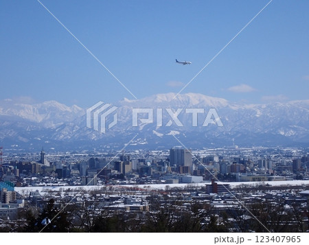 冠雪の北アルプス薬師岳を背景に雪景色の富山市街地と着陸態勢の旅客機 123407965