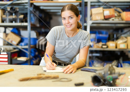 Smiling young woman storehouse worker standing at workbench and making notes Smiling young woman storehouse worker standing at workbench and making notes 123417293