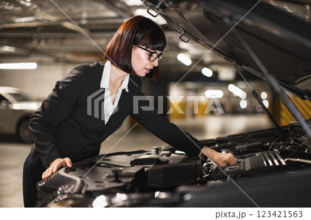 Caucasian businesswoman in formal attire fixes car engine in underground parking garage. Focused on repair, she wears glasses and stands by open hood, hands visible on engine. Caucasian businesswoman in formal attire fixes car engine in underground parking garage. Focused on repair, she wears glasses and stands by open hood, hands visible on engine. 123421563