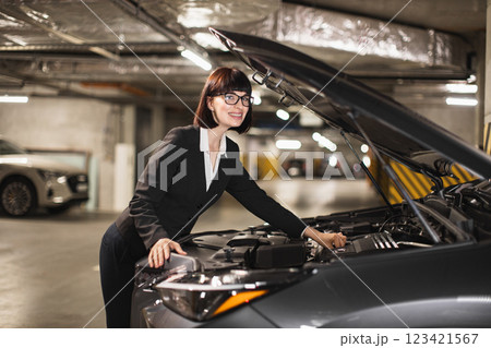 Caucasian businesswoman in formal attire opens car hood and inspects engine in underground parking. Scene conveys themes of independence and automotive repair skills. Caucasian businesswoman in formal attire opens car hood and inspects engine in underground parking. Scene conveys themes of independence and automotive repair skills. 123421567