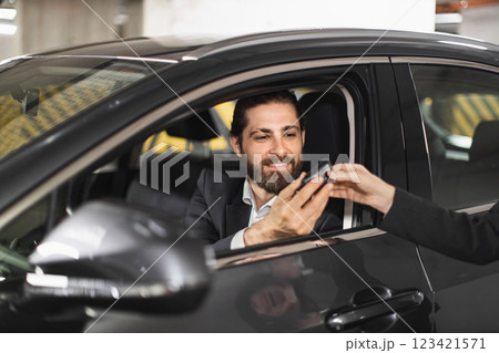 Caucasian businessman in suit receives car keys from another person in urban parking garage. Young adult male, beard, long hair, professional setting, modern vehicle. 123421571