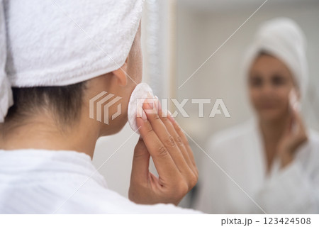 A close-up: A woman, towel-wrapped hair, applies a cleaning product to her face with a cotton pad while studying her reflection. A close-up: A woman, towel-wrapped hair, applies a cleaning product to her face with a cotton pad while studying her reflection. 123424508