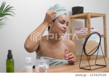 Tranquil beauty: A woman, with a towel-wrapped head, uses hyaluronic acid serum for her soothing skincare routine at the mirrored table. Tranquil beauty: A woman, with a towel-wrapped head, uses hyaluronic acid serum for her soothing skincare routine at the mirrored table. 123424526