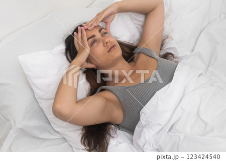 Frowning young lady, kneads temples, suffering a headache, reclined in a sunlit bed.  123424540