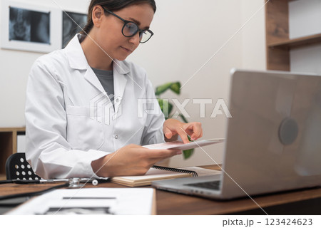 Medical Chart Examination: A female doctor in a white lab coat reviews patient medical documents while working at a table in a modern clinic. Medical Chart Examination: A female doctor in a white lab coat reviews patient medical documents while working at a table in a modern clinic. 123424623