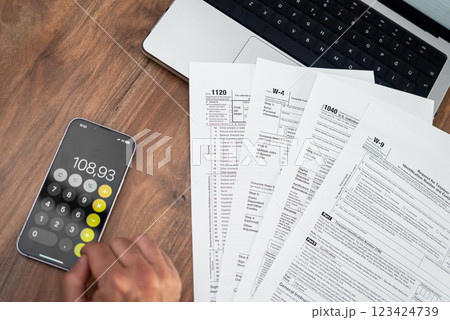 Person calculating taxes using a smartphone calculator with tax forms on a wooden desk. Financial management and tax filing concept. accountants, personal finance, and business tax preparation Person calculating taxes using a smartphone calculator with tax forms on a wooden desk. Financial management and tax filing concept. accountants, personal finance, and business tax preparation 123424739