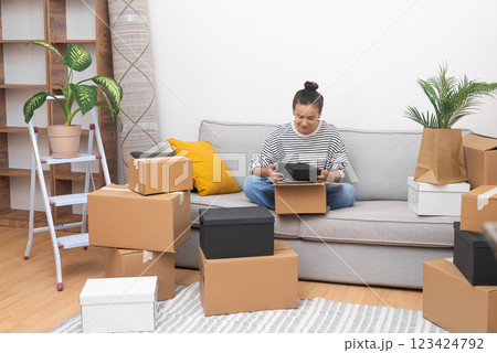 Smilling lady with bun o head with delighted expression sits on sofa collecting cardboard boxes for moving in new apartment with all modern furniture Smilling lady with bun o head with delighted expression sits on sofa collecting cardboard boxes for moving in new apartment with all modern furniture 123424792